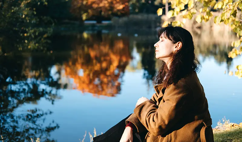 Mujer junto a un lago en otoño, con los ojos cerrados, conectando con lo que siente